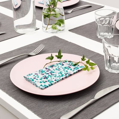 Table setting with pink plates, colourful pencils napkins, silverware, and glass cups. Greenery in vases adds decor.