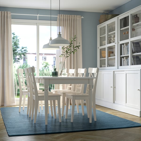 White dining table with 8 chairs, blue rug, pendant lamp. Blue room, large windows.