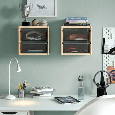 TROFAST shelves with dark boxes hold sports items & books. Desk below has a lamp, books, pens, tablet, water bottle against a light blue wall.