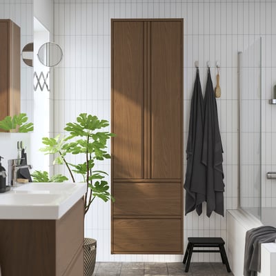 Modern bathroom with tall brown oak storage cabinet, drawers, and doors. Plant and dark towels nearby.