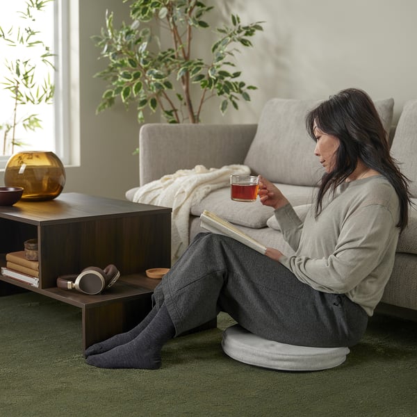 Person reads book on sofa, sipping tea using ÅKERVINDEFLY memory foam seat cushion.