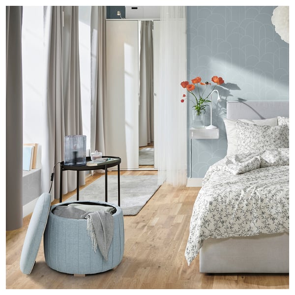 Blue patterned bedroom with wooden floor and large window. Floral bed, round ottoman, side table with poppies and candle.