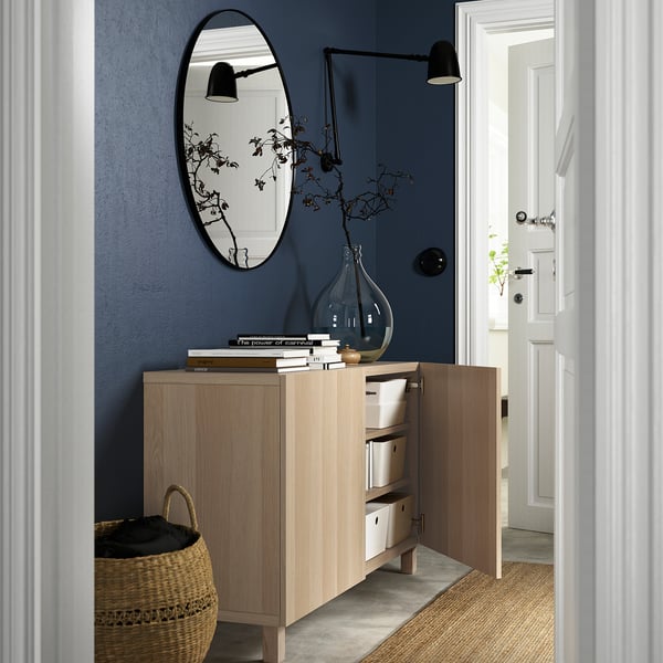 Hallway with light wood BESTÅ sideboard, open doors revealing storage boxes. Above, round mirror and wall lamp on blue wall.