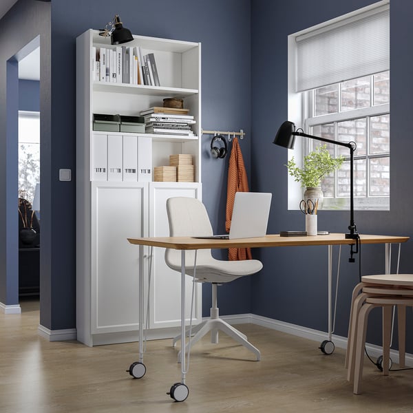 Modern home office with a white BILLY bookcase filled with books and decor, a wooden desk, grey chair, and a window with blinds.