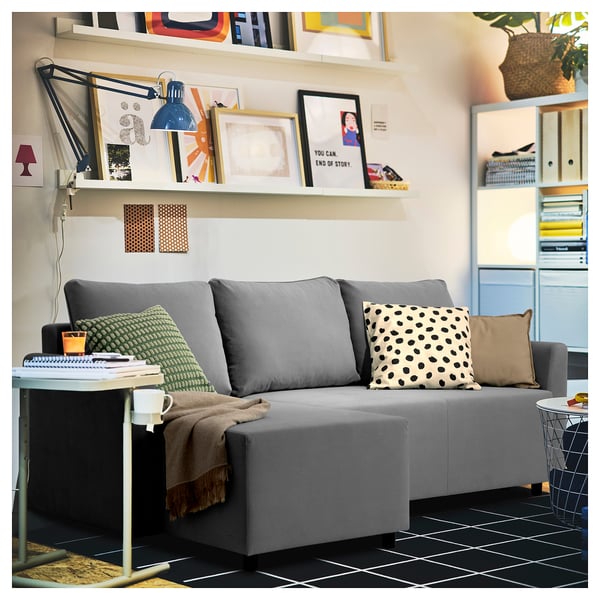 Modern grey l-shaped sofa with colourful cushions and throw, under shelf with books and lamp, black-tiled floor.