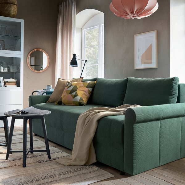 A cosy living area with a DÅNHULT green sofa bed, decorated with patterned pillows, situated near a window. A small side table holds a mug and book.