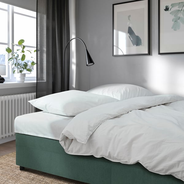 Modern minimalist bedroom with grey walls, large window, green sofa bed, white bedding, and lamp.