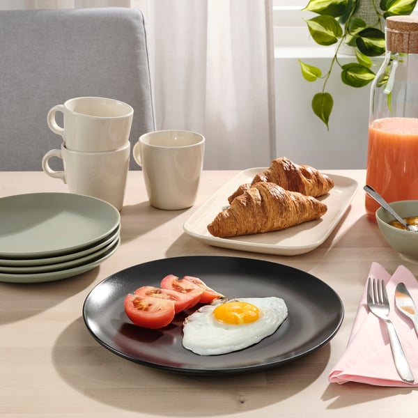 Plate with fried egg and tomatoes, stacked mugs, croissants on tray, juice, and cutlery on table.
