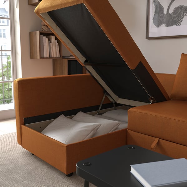 An orange sofa bed with a lift-up chaise section revealing storage space with pillows inside.