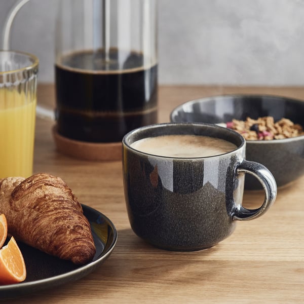 Dark grey mug, orange juice, croissant, and granola on a wooden table, showcasing a stylish breakfast setup.