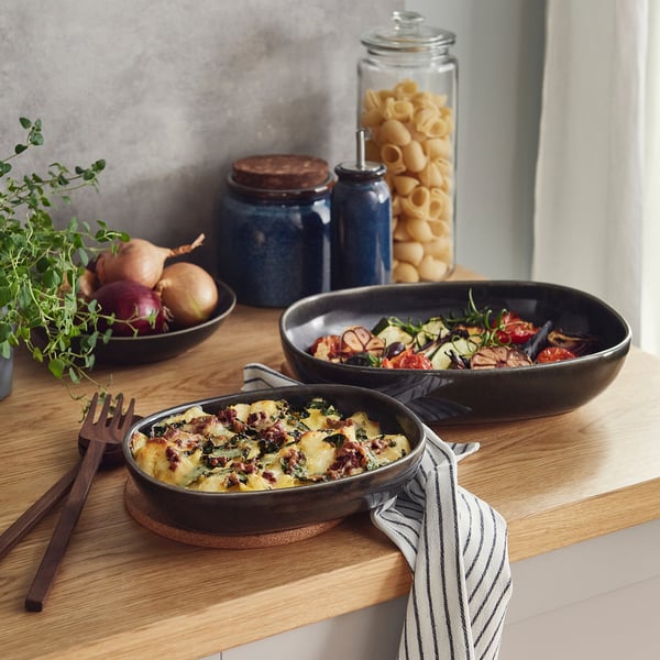 Kitchen counter with two glazed oven dishes: one with vegetable casserole, one with roasted veg. Surrounded by utensils, herbs, and kitchenware.