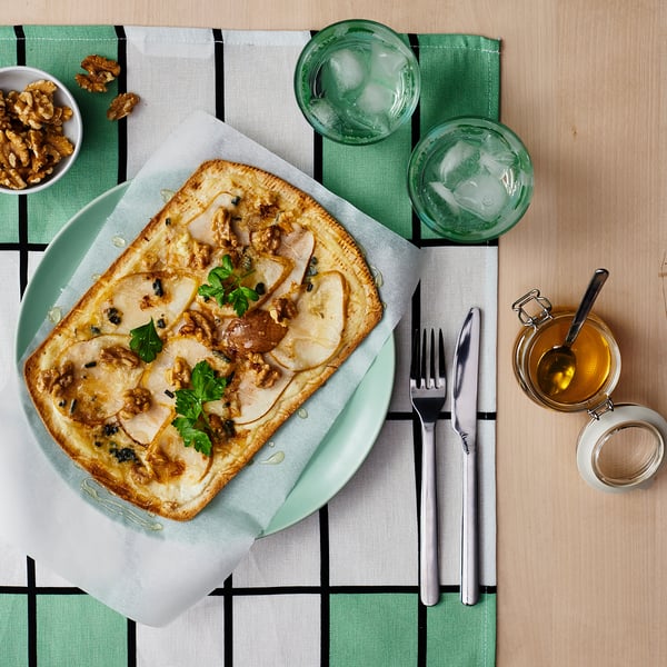 A green plate with flatbread topped with cheese, nuts, and herbs accompanied by glasses of water and a bowl of nuts on a green-plaid tablecloth.