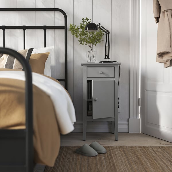 Bedroom scene with black metal bed, grey GULLABERG bedside table, lamp, plant, and slippers.