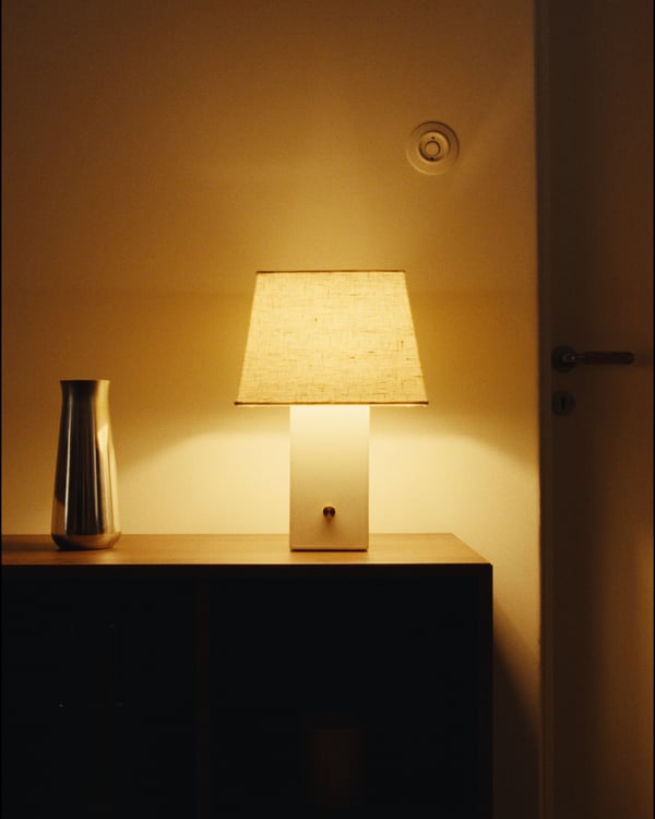 A HALGATT lamp with a beige linen shade and white ceramic base on a wooden surface, next to a silver vase.