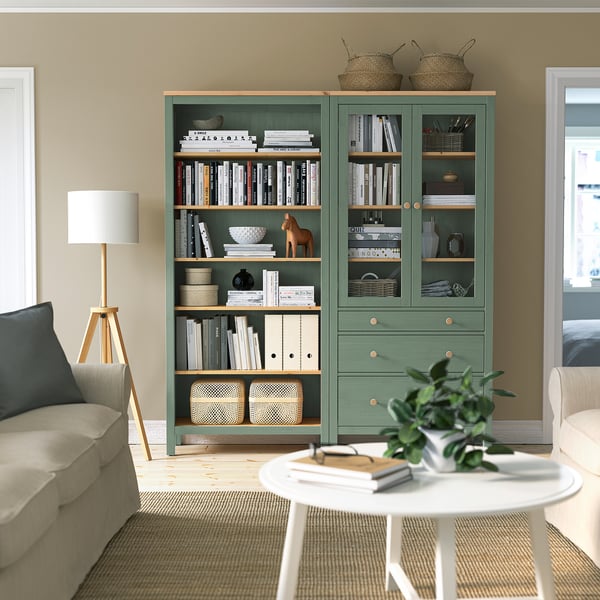 Cosy living area with large green HEMNES bookcase, wooden floor lamp, beige rug, and cream couch. Books and decor items displayed neatly on shelves.