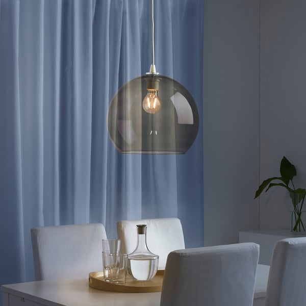 Modern dining setup with smoked glass light, white chairs, clear glasses, wooden tray, and vase. Curtains drawn.