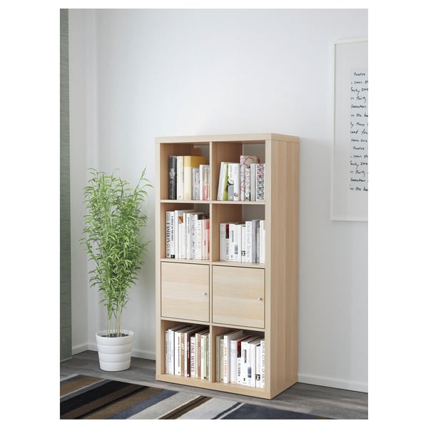 Light wood KALLAX shelf, holding books, against a wall. Accompanied by tall plant.