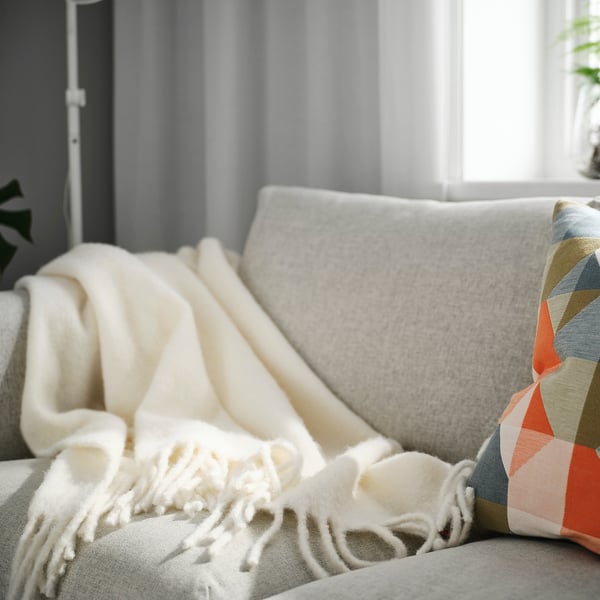 Off-white cosy throw blanket with fringe, larger than cushion, on cream sofa.