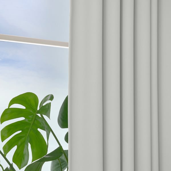 Window with white block-out KORGMOTT curtains showing pleats, providing privacy and light control. Large green plant partially visible.