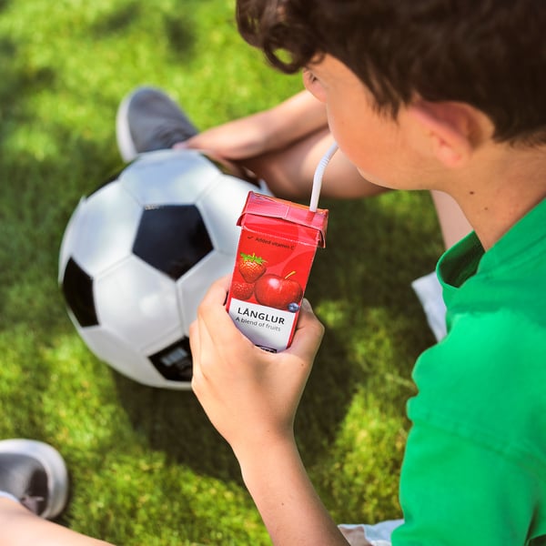 A child drinks LÅNGLUR fruit smoothie on grass near a football ball, green shirt.