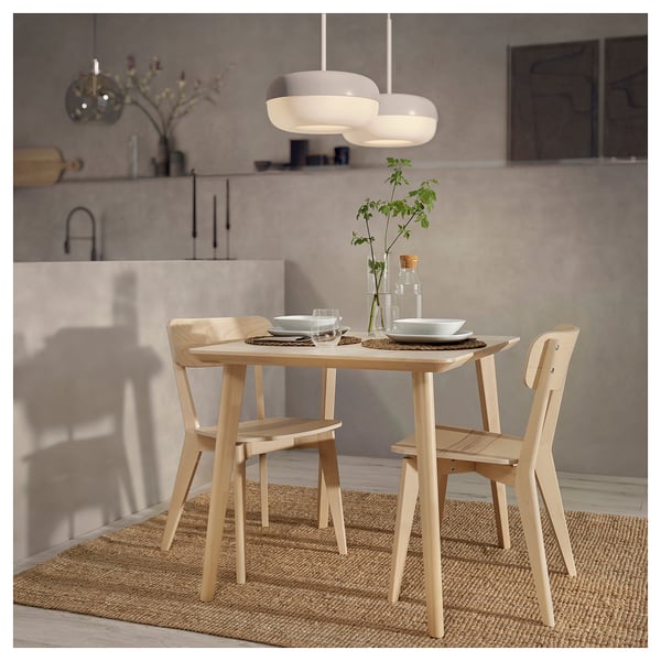 Modern dining set with light wood table and chairs, hanging light, plates, glasses, and plant.