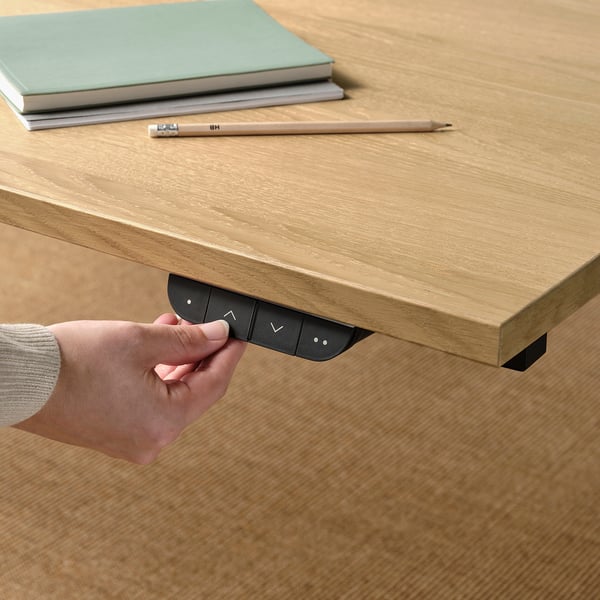 A hand adjusts a sit-stand desks control panel, with a wooden top holding books and a pencil.