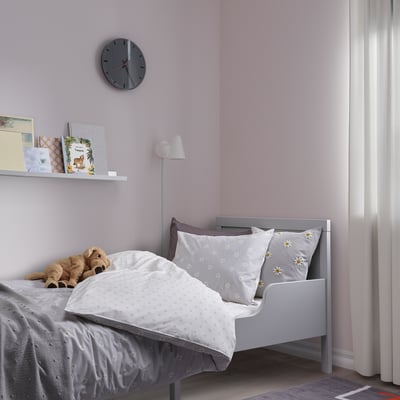 A cosy NATTSLÄNDA daisy-print grey and white duvet cover, made of cotton, neatly fits a small bed in a pink room. A wall clock and books are above it.