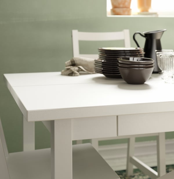 White wooden dining table with dark plates, bowls, and jug. Green wall background.