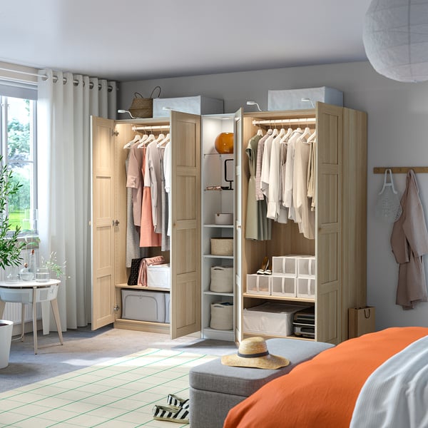 Light-filled room with open wooden wardrobe, PAX, displaying hanging clothes, shelves with folded clothes, and storage boxes.