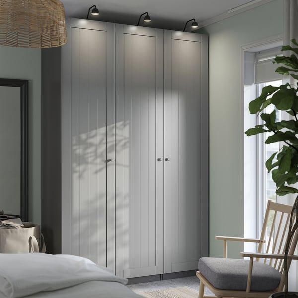 Modern bedroom with tall grey PAX wardrobes, lit tops, white bed, wooden chair, and large plant by the window.
