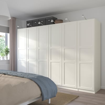 Large white PAX wardrobe with shelves/drawers, next to bed with blue/beige bedding. Minimalist design, lit overhead. Larger than plant, bed.