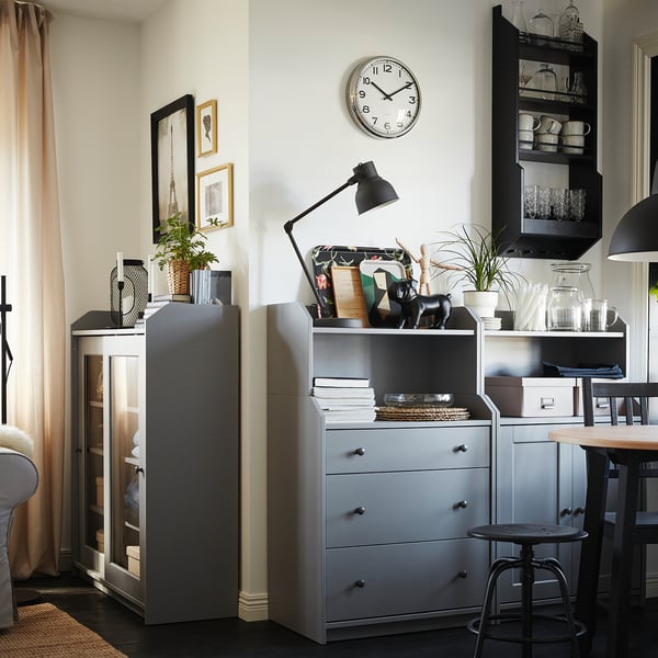 Modern living room with gray cabinets, a round wall clock, black lamp, and decorative items, with a dining table and chairs in the background.