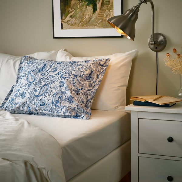 Bedroom with RODGERSIA paisley pillowcases, beige bedding, books, dried flowers, warm lamp.