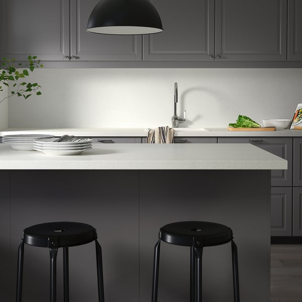Modern kitchen counters with contrasting light and dark tones; black round stools neatly positioned; clean plates and utensils on the counter.