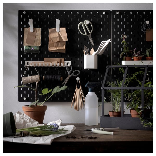 Black SKÅDIS pegboard with various kitchen tools and plants attached, including scissors, bags, and spray bottle, arranged against a wall.