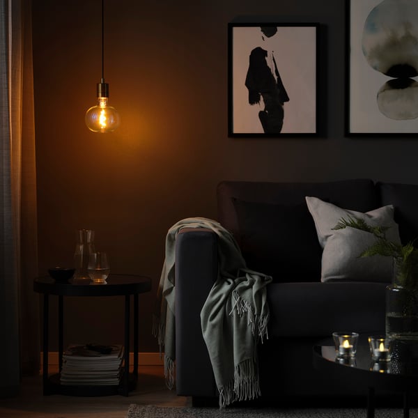 Dim room with SKAFTET lamp over table. Grey sofa with blanket, pillows, and wall art.