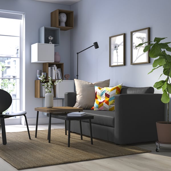 Modern living room with grey sofa bed, colourful pillows, wooden coffee table, and plants.