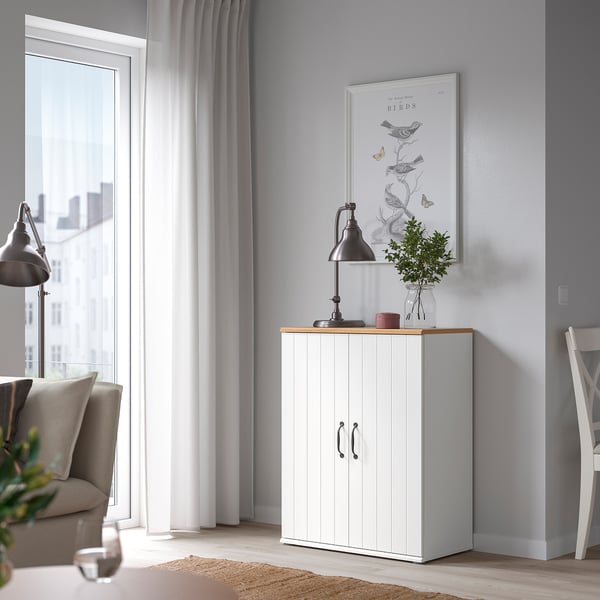 A cosy living room featuring a white SKRUVBY cabinet with double doors, oak top, and black decor. Balanced with a window and chair.