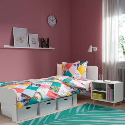 Bright pink kids room with white bedside table, yellow box, books, colourful bedspread, and striped green rug.