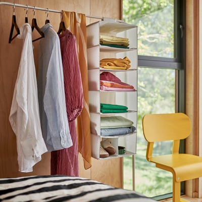 A bedroom scene with a striped bedsheet, two hangers holding clothes, and a yellow chair beside a window.