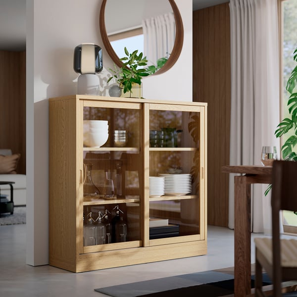 TONSTAD light oak cabinet, glass doors, three shelves. Stores dishes, bottles. Shows classic design. Tall, fits well in room with other furniture.