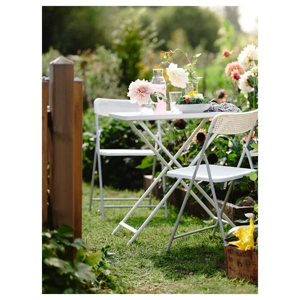 Small outdoor dining set up with white metal table and chairs, flowers and glasses, situated on grass.