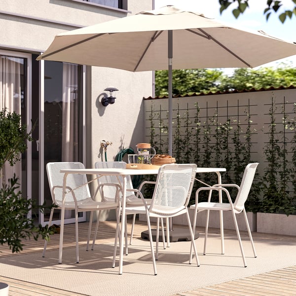 Modern outdoor patio with white TORPARÖ table and chairs, beige umbrella, set on wooden deck, surrounded by greenery.