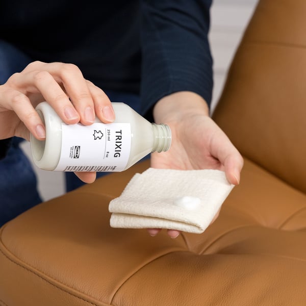 Person applies leather conditioner from a bottle labelled TRIXIG onto a cloth, preparing to clean and protect leather furniture.