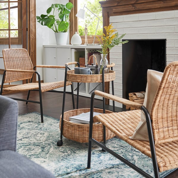 Cosy living room with rattan chairs, woven table, and patterned rug. Brick fireplace and large windows.