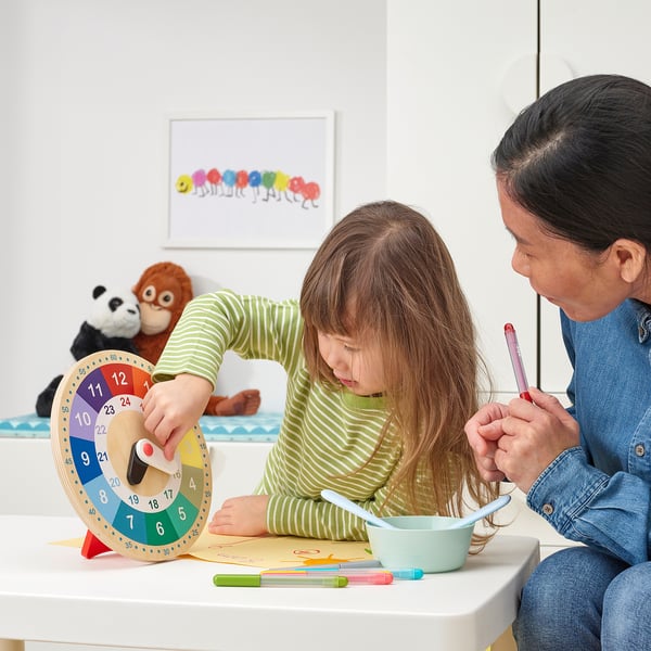 Child and adult sit at table, using educational clock and colouring. Clocks hands are moved, showing learning time.