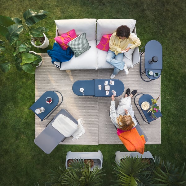 Two people relax outdoors on foldable VIGGSÖ lounge chairs; one plays cards while the other reads, surrounded by greenery.