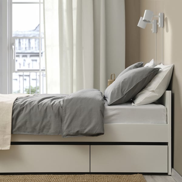 A modern bed with two storage drawers, grey bedding, placed near a window with light curtains. A wall light and a rug are visible nearby.
