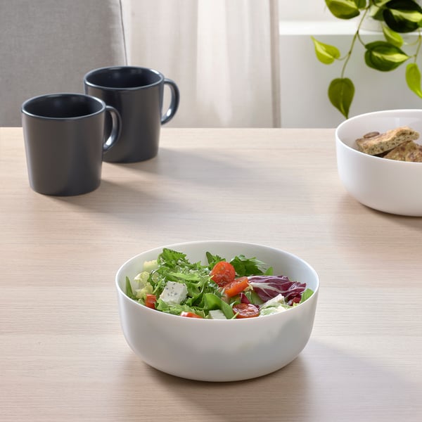 Two white bowls on a table, one with salad, one with bread. Two black mugs nearby. Bowls are wide and shallow, made of tempered glass.
