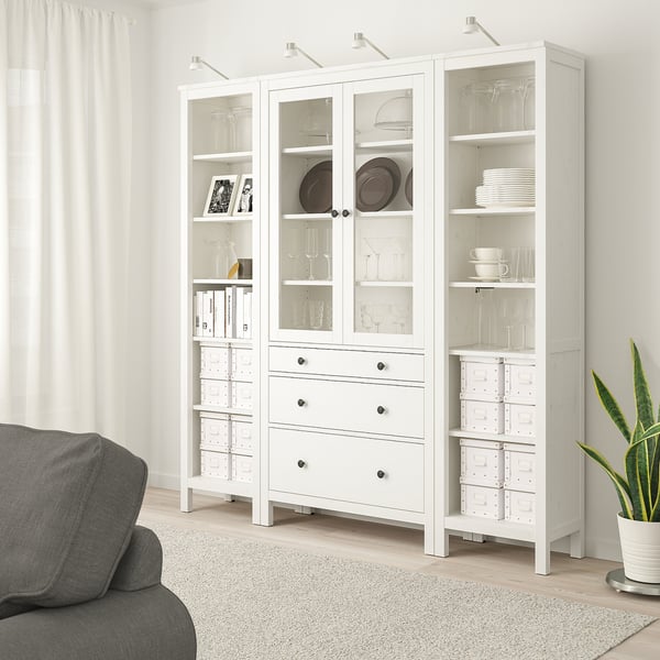 Белый шкаф HEMNES со стеклянными дверцами, полками и ящиками рядом с серым диваном, где выставлена стеклянная посуда, посуда и книги.